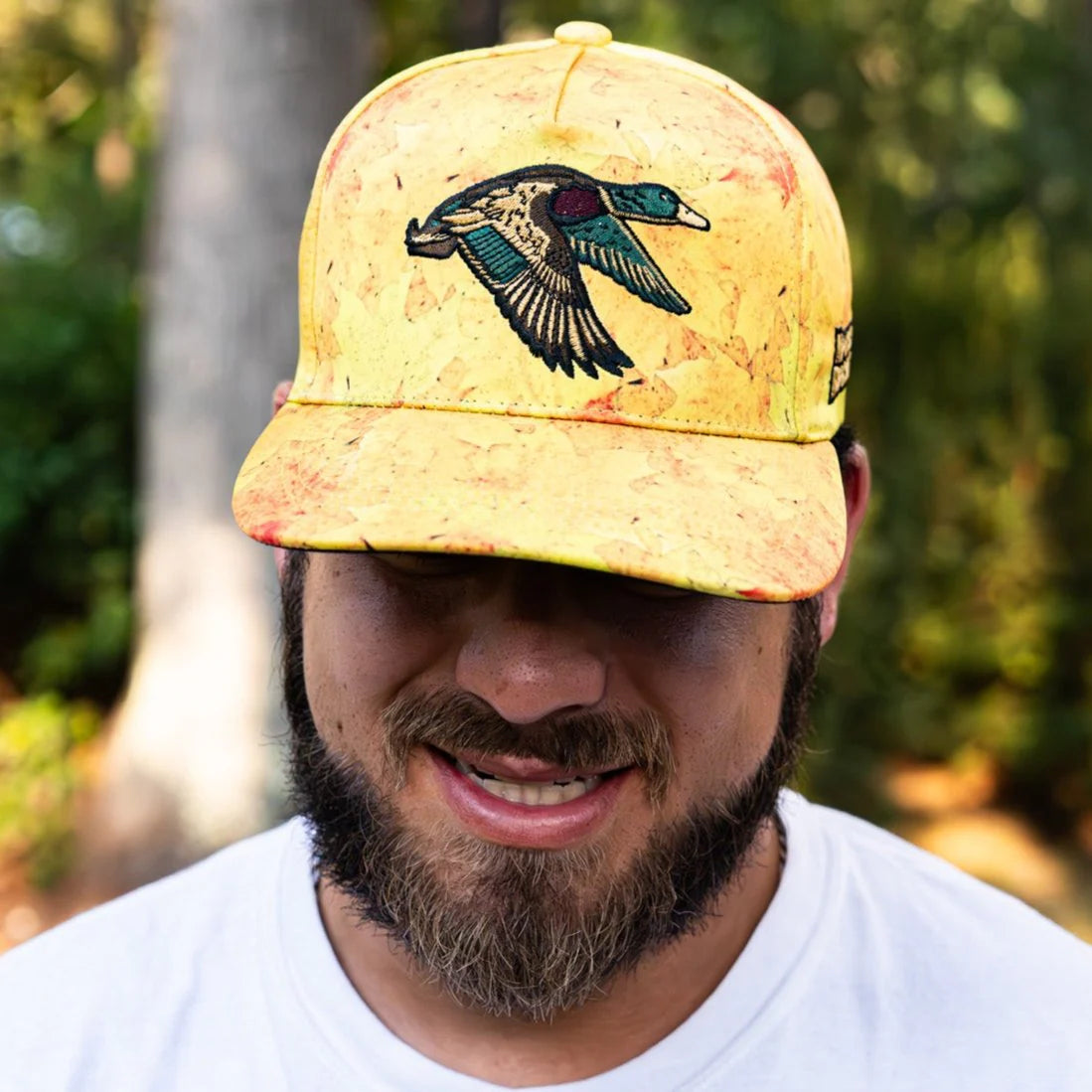 A bearded man stands outdoors, smiling beneath a Golden Leaf Camo Duck Bucket from Mucker Bucket. The blurred, lush green background enhances his relaxed demeanor.