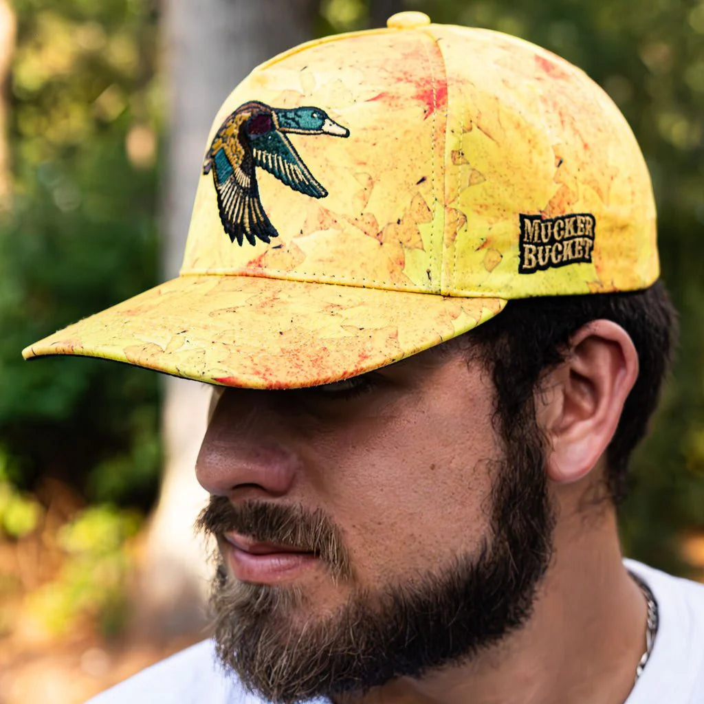 A bearded man wears the Golden Leaf Camo Duck Bucket hat by Mucker Bucket, with a bird design and logo, reminiscent of duck hunting adventures. Blurred green foliage forms his backdrop.
