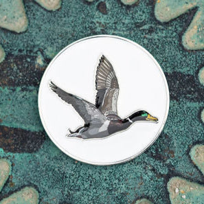 The OG Duck Lucky Huntin Coin pin by Mucker Bucket features a mallard duck in flight, detailed against a white background with realistic colors on a textured green surface, making it a must-have for collectors.