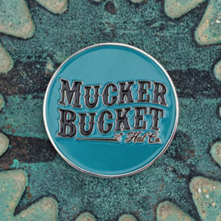 A round teal pin adorned with Mucker Bucket in bold black letters against a textured background with irregular shapes and a subtle duck design, named Duck, Duck, Boom Lucky Huntin Coin.