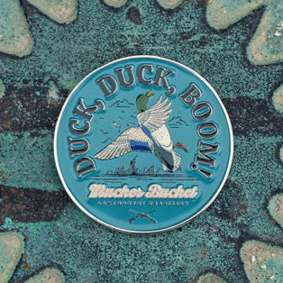 Round pin badge featuring a flying duck and the text Duck, Duck, Boom! with Winchester Buckshot below. The teal background adds texture, reminiscent of Mucker Buckets Duck, Duck, Boom Lucky Huntin Coin.