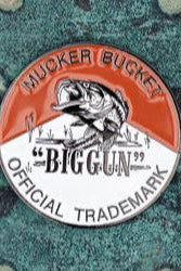 The Biggun Lucky Fishin Coin by Mucker Bucket features a circular logo with a central fish illustration. Mucker Bucket arches at the top, with Official Trademark below, all set against a textured, rusted metal background reflecting fishing tales and coin lore.