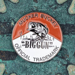 The Biggun Lucky Fishin Coin by Mucker Bucket features a circular logo with a central fish illustration. Mucker Bucket arches at the top, with Official Trademark below, all set against a textured, rusted metal background reflecting fishing tales and coin lore.