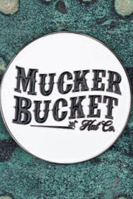 A round pin bearing the text Mucker Bucket rests on a textured teal and rust surface, evoking the charm of a Biggun Lucky Fishin Coin, poised to bring luck.