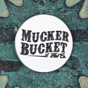 A round pin bearing the text Mucker Bucket rests on a textured teal and rust surface, evoking the charm of a Biggun Lucky Fishin Coin, poised to bring luck.