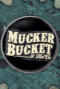 The Aim Small, Miss Small Lucky Huntin Coin by Mucker Bucket, a round black pin with Mucker Bucket Hat Co. in white text, rests on a textured teal and gray surface, reminiscent of an intricate hunting pattern for those blending style and stealth.