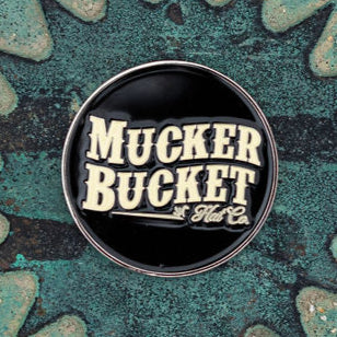 The Aim Small, Miss Small Lucky Huntin Coin by Mucker Bucket, a round black pin with Mucker Bucket Hat Co. in white text, rests on a textured teal and gray surface, reminiscent of an intricate hunting pattern for those blending style and stealth.