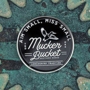 A round black sticker with white text reads Mucker Bucket Southern Goods, featuring an illustration of a duck above it, evocative of the Aim Small, Miss Small Lucky Huntin Coin. The background showcases star-like patterns on a textured greenish-blue surface.