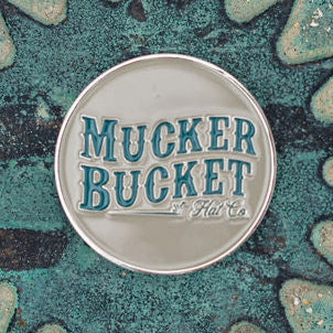 A round metal emblem with raised blue-green Mucker Bucket Hat Co. lettering on a silver background resembles the Chasin Tail Lucky Huntin Coin from Mucker Bucket. It rests on a textured, weathered green surface.