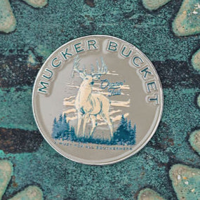 The Chasin Tail Lucky Huntin Coin by Mucker Bucket features a stag in mountains and trees. With Crown Tail and Made in the Netherlands on it, set against a speckled green and brown background, its perfect for your next adventure.