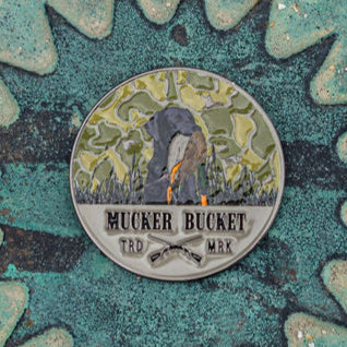 The Got One Lucky Huntin Coin is a round metal pin with a person shoveling, crossed shovels, and Mucker Bucket TRD MRK on it. Its green camouflage background hints at adventure, making it a lucky charm for any bold collector.