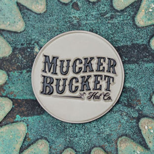 A gray patch with bold black Mucker Bucket Hat Co. text rests on a textured teal backdrop with gear patterns, resembling the Got One Lucky Huntin Coin. Why not get one for yourself?.