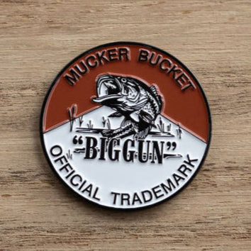 A circular emblem with a fish leaping from water resembles the Biggun Lucky Fishin Coin. The top half displays the Mucker Bucket brand in brown, while the bottom half is white, featuring the bold Biggun Official Trademark. The background has a wooden texture.
