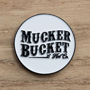 A round pin with Mucker Bucket in bold black text on a white background rests on wood, like the Biggun Lucky Fishin Coin washed ashore, awaiting adventures.