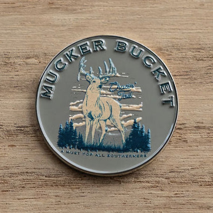 The Mucker Bucket Chasin Tail Lucky Huntin Coin features a round metal pin with a detailed buck, antlers, trees, and clouds. The text A Must for All Southerners rests on a wooden backdrop, embodying the Southern spirit.