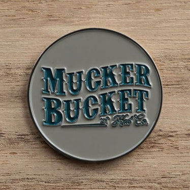 A round metallic badge embossed with Mucker Bucket in stylized blue-green lettering against a rustic wooden backdrop, like the Chasin Tail Lucky Huntin Coin, ready to bring fortune on your next hunt.
