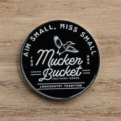The Aim Small, Miss Small Lucky Huntin Coin by Mucker Bucket features a round black design with white text and a flying duck illustration. Below, it reads, A Lowcountry Tradition. This unique piece is an ideal addition to any hunting enthusiasts collection.