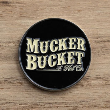 A round, black pin with Mucker Bucket in bold, yellow letters rests on a wooden surface, much like the Aim Small, Miss Small Lucky Huntin Coin waiting to be found.