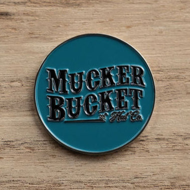 A circular teal metal pin embossed with Mucker Bucket Hat Co. lies on a wooden surface next to the Duck, Duck, Boom Lucky Huntin Coin charm.