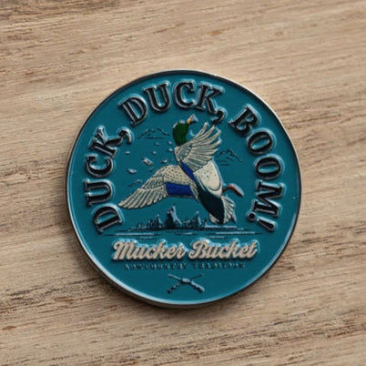 A round blue pin badge features a flying duck with DUCK, DUCK, BOOM! above and Mucker Bucket below, reminiscent of the charm of the Mucker Bucket brands Duck, Duck, Boom Lucky Huntin Coin.
