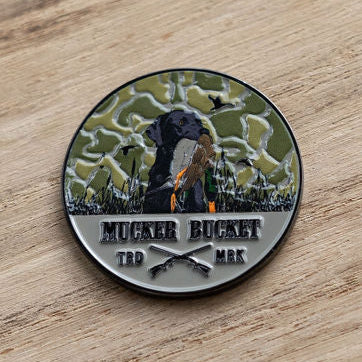 The Got One Lucky Huntin Coin by Mucker Bucket features a black dog with a bird in its mouth on a camo background. It says MUCKER BUCKET with crossed rifles below and TRD MRK on the sides, all resting on a wooden surface, capturing the essence of luck in hunting.