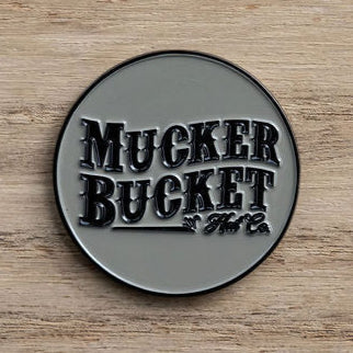 This Got One Lucky Huntin Coin by Mucker Bucket is a round metal badge featuring embossed letters against a wooden backdrop with a glossy finish, exuding charm and serendipity.