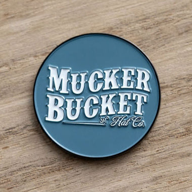 A blue, round pin sits on a wooden surface, featuring white text with Mucker Bucket branding in a vintage style, echoing the Smart Duck Lucky Huntin Coin era.
