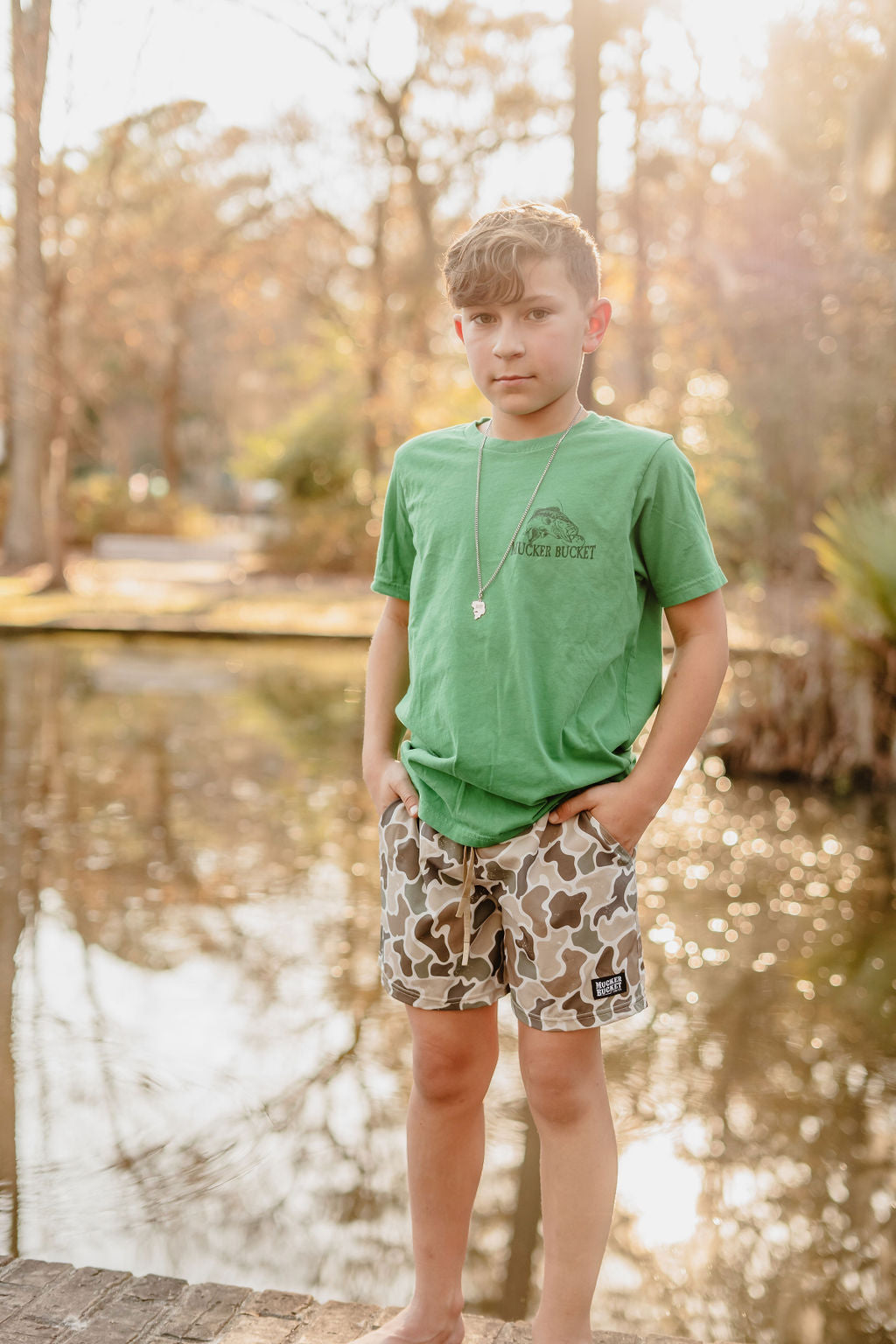 A young boy stands barefoot by a pond, wearing a green t-shirt and Mucker Buckets Youth Performance Athletic Shorts in camo. His hands are in his pockets, with a necklace around his neck. The sunlit forest background is softly focused, adding a warm glow to the scene.
