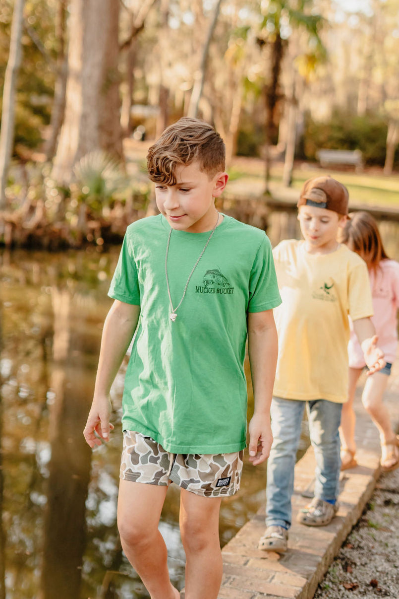 A boy in Mucker Buckets Youth Catch of the Day shirt and camo shorts wanders along a pond trail. Hes with kids—one in a yellow cap and another in pink, surrounded by trees on their scenic adventure wearing youth-sized outfits.