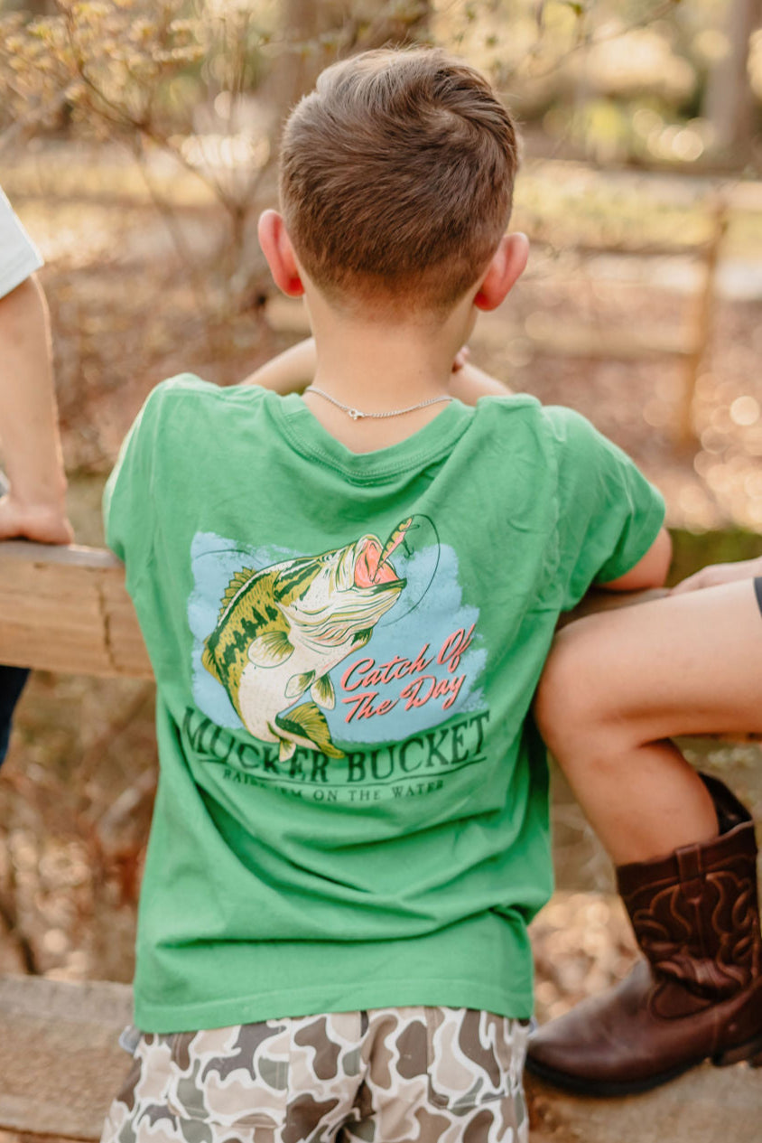 A young boy in a Youth Catch of the Day tee by Mucker Bucket, featuring a fish graphic, stands on a wooden bench outside. Children around him wear cowboy boots and camouflage shorts, enjoying the sunny, wooded setting.