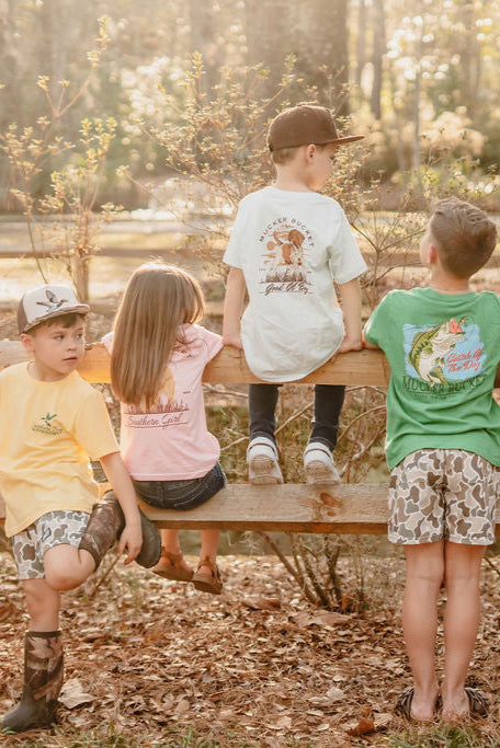 Four children outside, seated and standing on a wooden bench, wear Mucker Buckets Youth Good Ol Boy GSP tees featuring playful dog graphics. In the sunlit, leafy area, warm natural light filters through the trees as they enjoy their day in vibrant country styles from sizes XS-XL.
