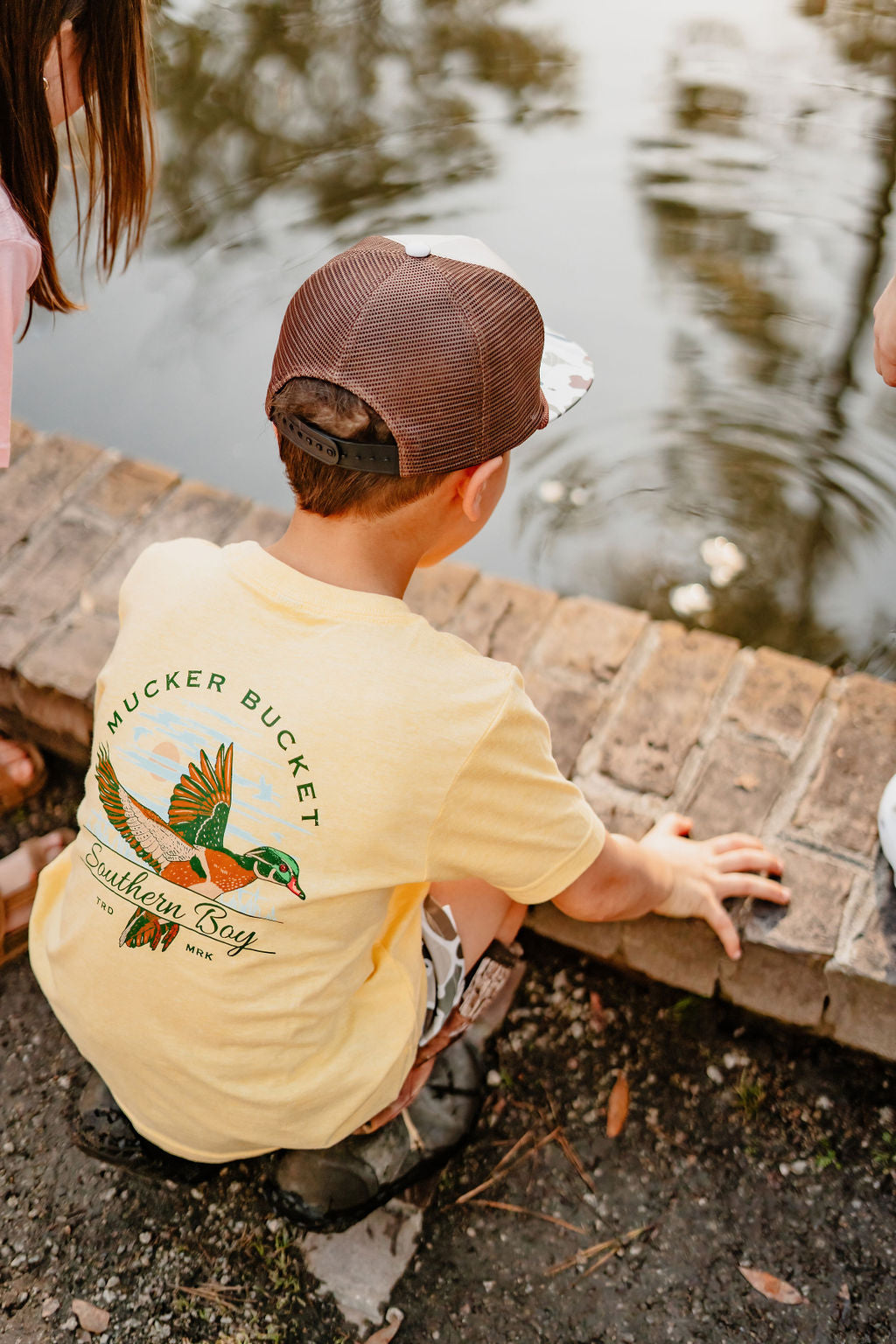 A child in a yellow Youth Southern Boy tee by Mucker Bucket, with a flying duck graphic, crouches by the pond as ripples dance on the water. A brown cap shields their eyes while two other children are partially visible nearby.