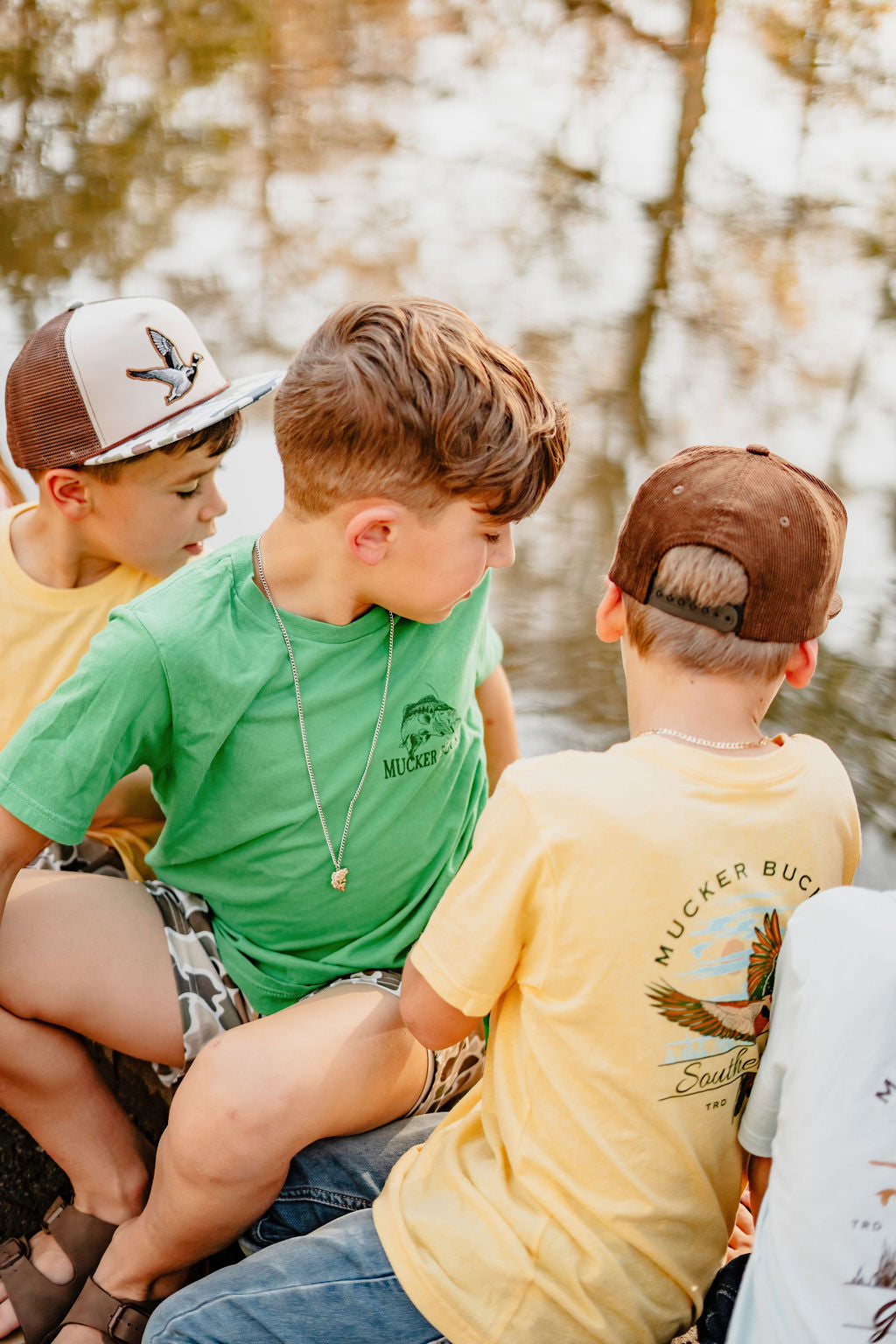 Three children sit by a pond in country-style attire with caps. One wears a green shirt and camo shorts, another sports Mucker Buckets Youth Catch of the Day yellow tee featuring a fish graphic, while the third wears white. They gaze at the water reflecting tree images.