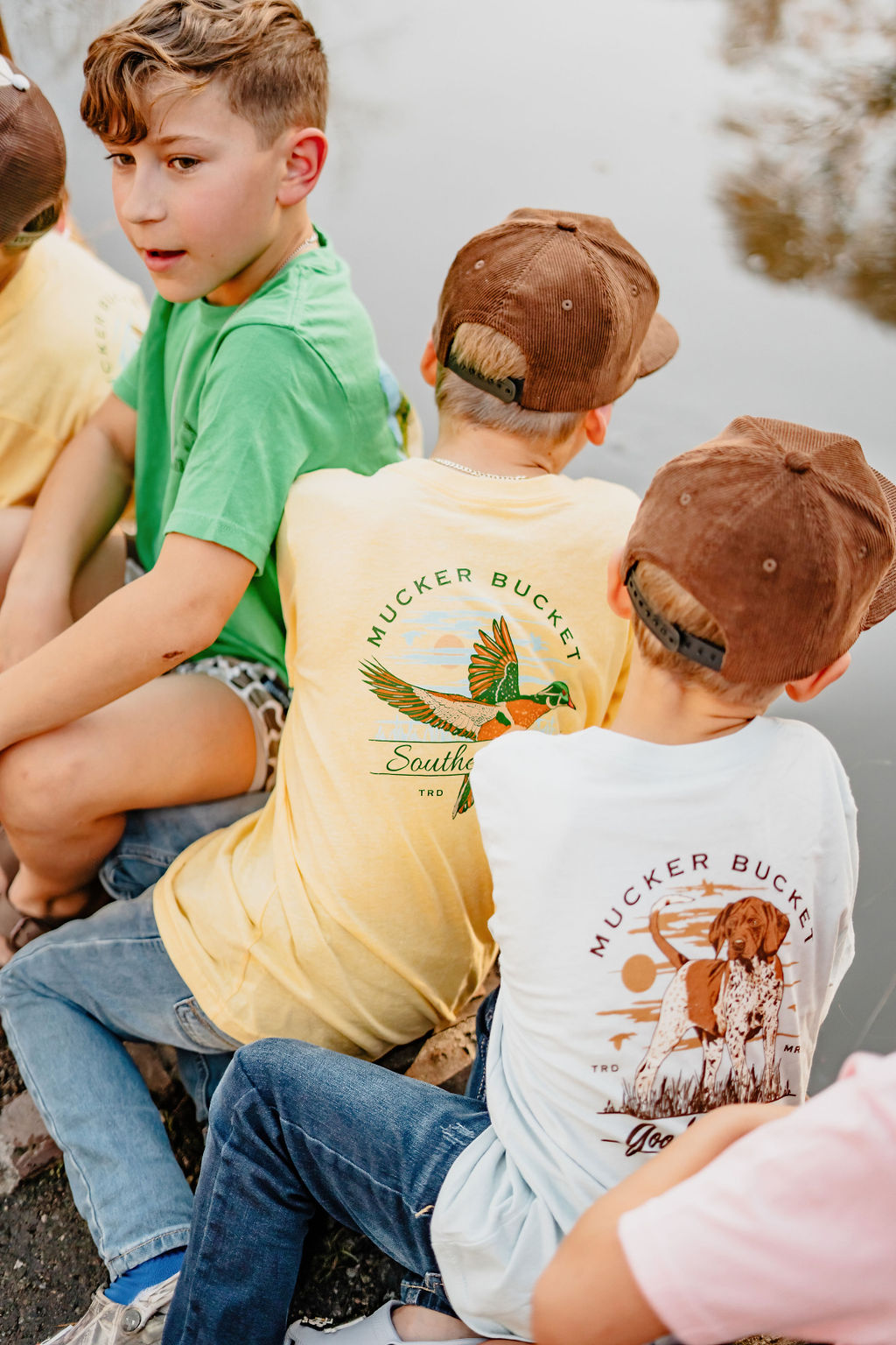 A group of children in casual clothes and brown caps sits by a body of water. One wears a yellow shirt with a parrot design, while another sports the Mucker Bucket Youth Good Ol Boy GSP country-style tee featuring a cowgirl illustration. Available in youth sizes XS-XL.