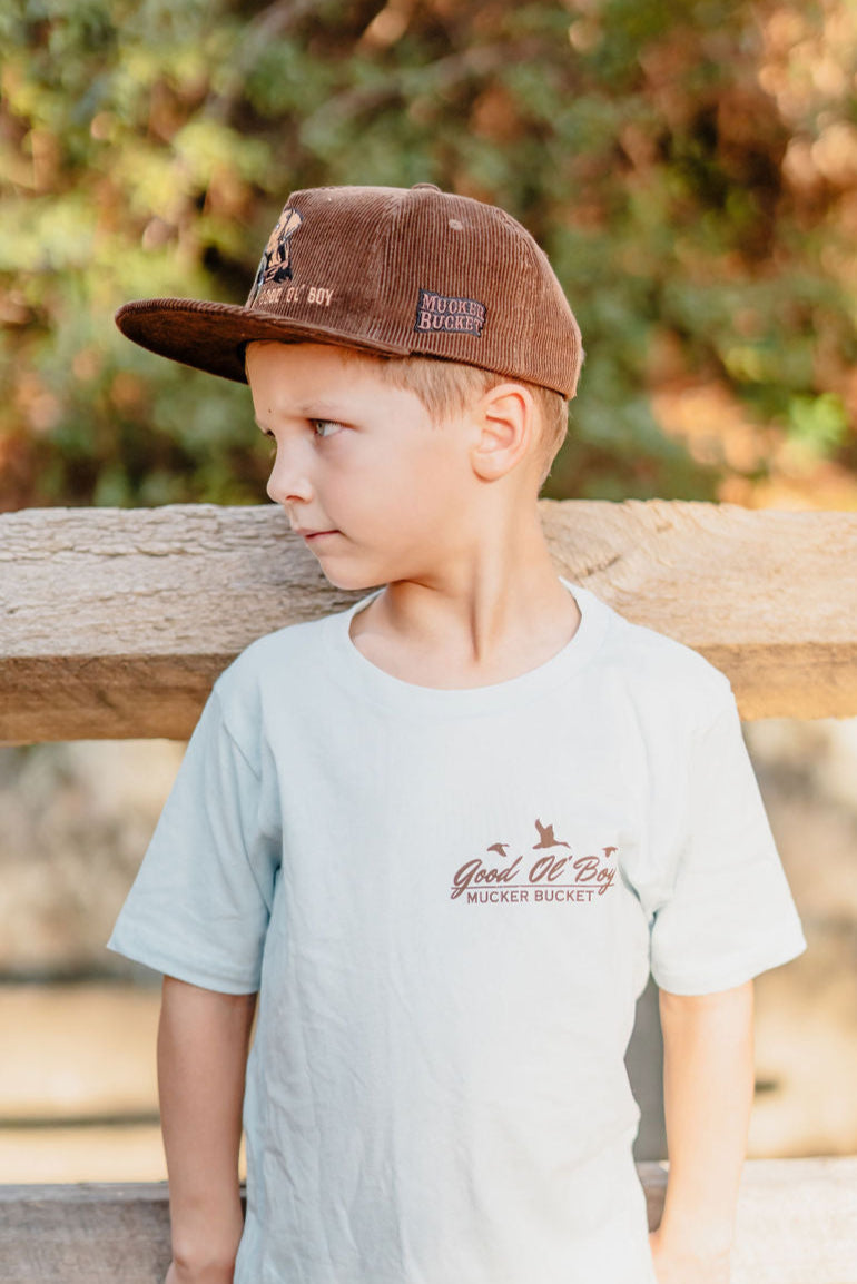 A boy wearing the Mucker Bucket Youth Good Ol Boy GSP tee stands near a wooden fence glancing right. His brown cap complements the scene, with a blurred backdrop of lush green foliage.