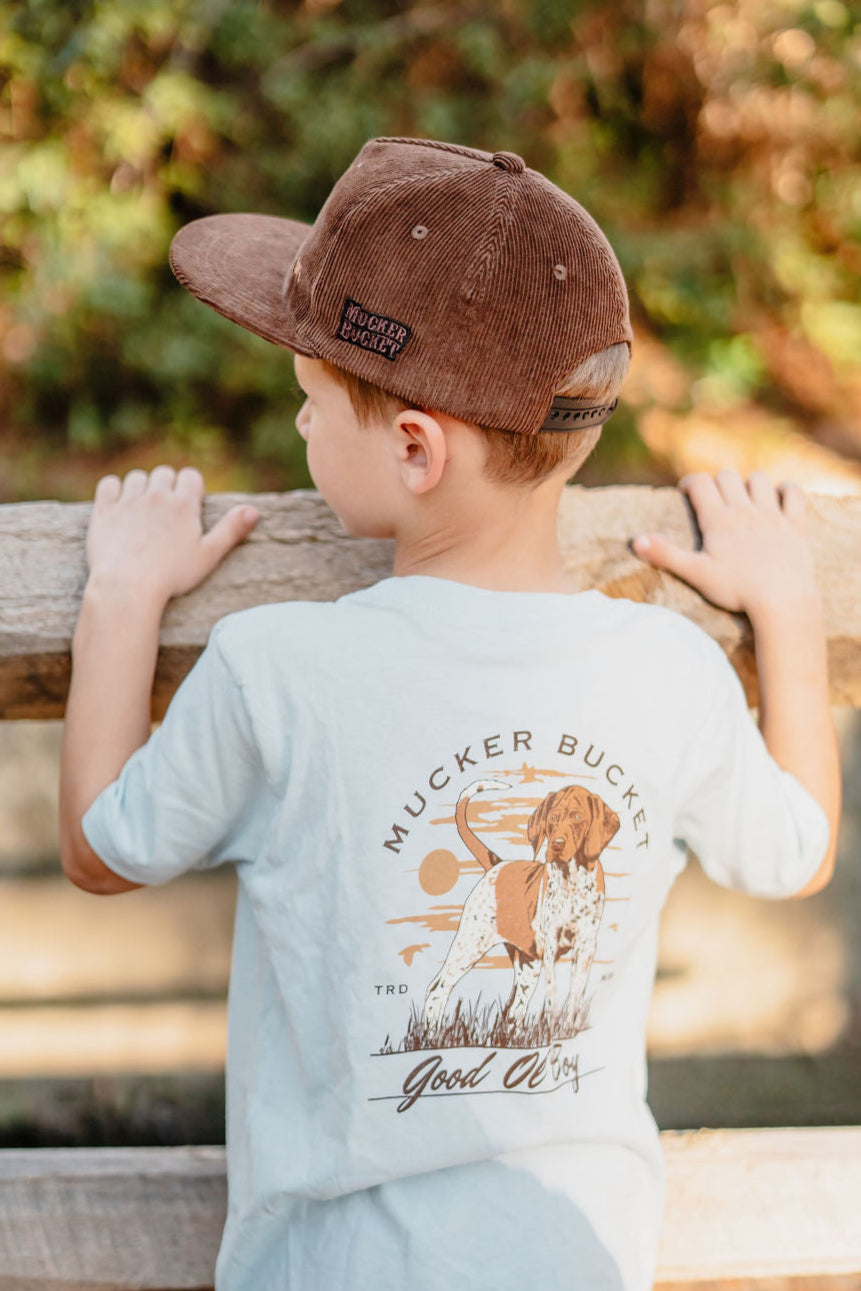 A young boy in a brown cap wears a light blue Mucker Bucket Youth Good Ol Boy GSP tee with a dog graphic. He stands by a wooden fence, embodying the charm of this country-style shirt, available in youth sizes XS-XL, with softly blurred bushes in the background.
