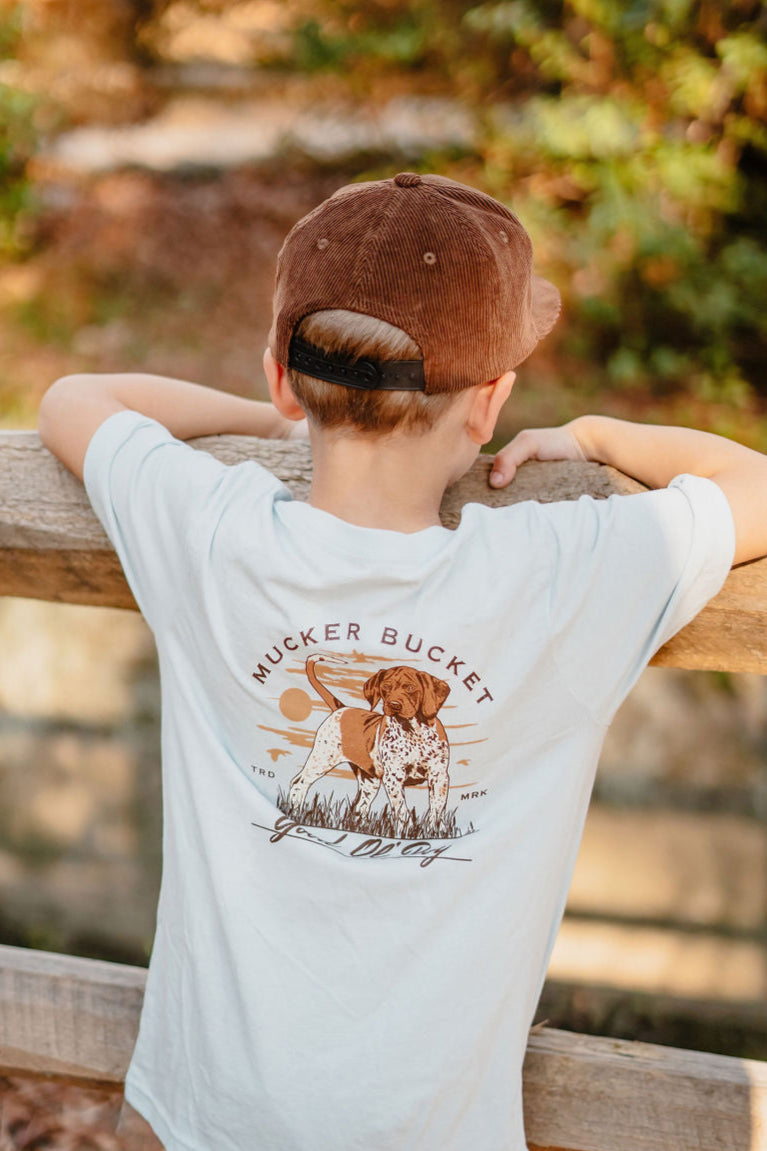A boy in a brown cap leans on a wooden fence, wearing the Youth Good Ol Boy GSP tee by Mucker Bucket, featuring an illustrated dog and Mucker Bucket text. The white T-shirt, available in youth sizes XS-XL, stands out against the blurred greenery background.