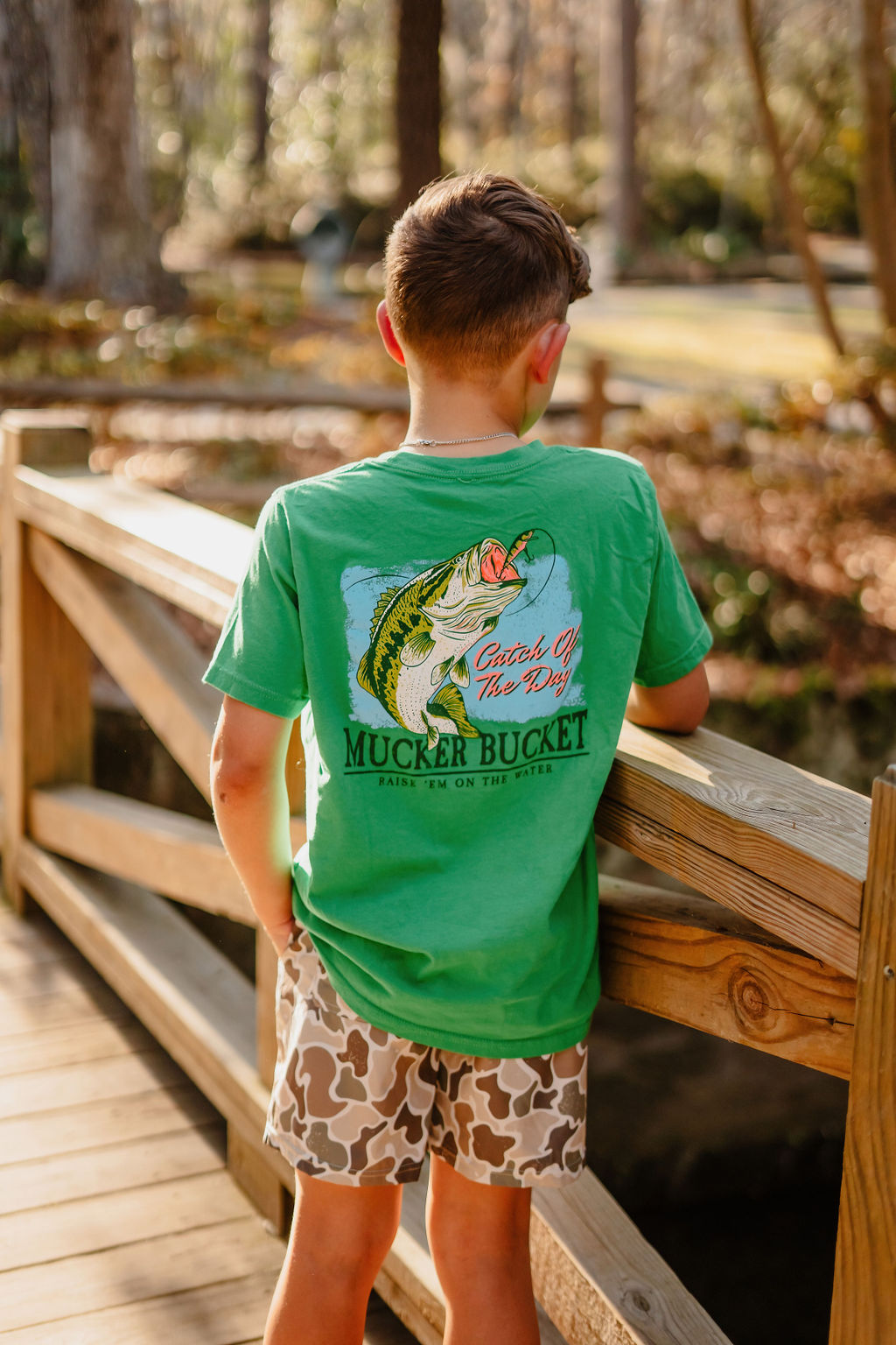 A boy wearing the Mucker Bucket Youth Catch of the Day green T-shirt and camo shorts stands on a wooden bridge, facing away. The tee showcases a fish graphic and phrases like Catch of the Day and Mucker Bucket, with trees and a creek as a scenic backdrop, ideal for exploration in youth sizes.