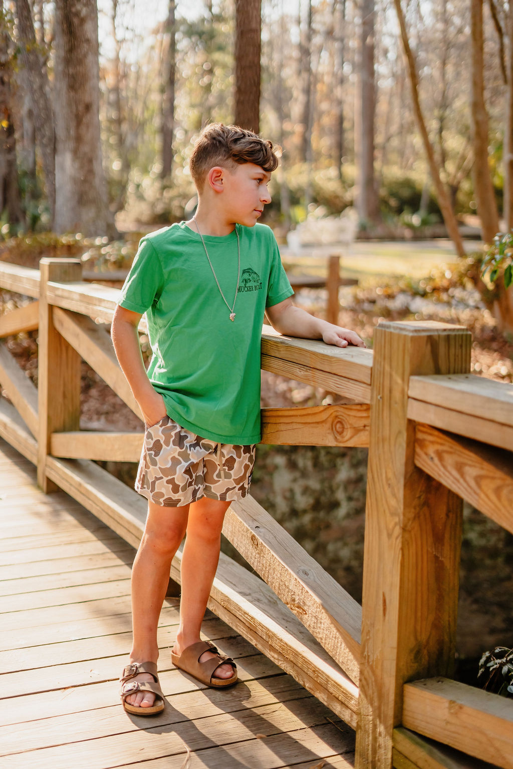 A boy in a green T-shirt and Mucker Buckets Duck It Youth Performance Athletic Shorts stands on a wooden bridge in a sunny forest, gazing sideways amidst lush greenery.