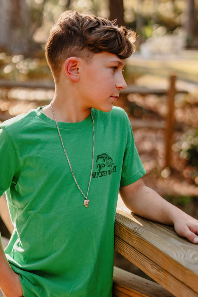 A boy in a Mucker Bucket Youth Catch of the Day country-style green tee leans against a wooden railing outdoors. Wearing a silver necklace, he gazes to the side amidst blurred greenery and trees in the background.