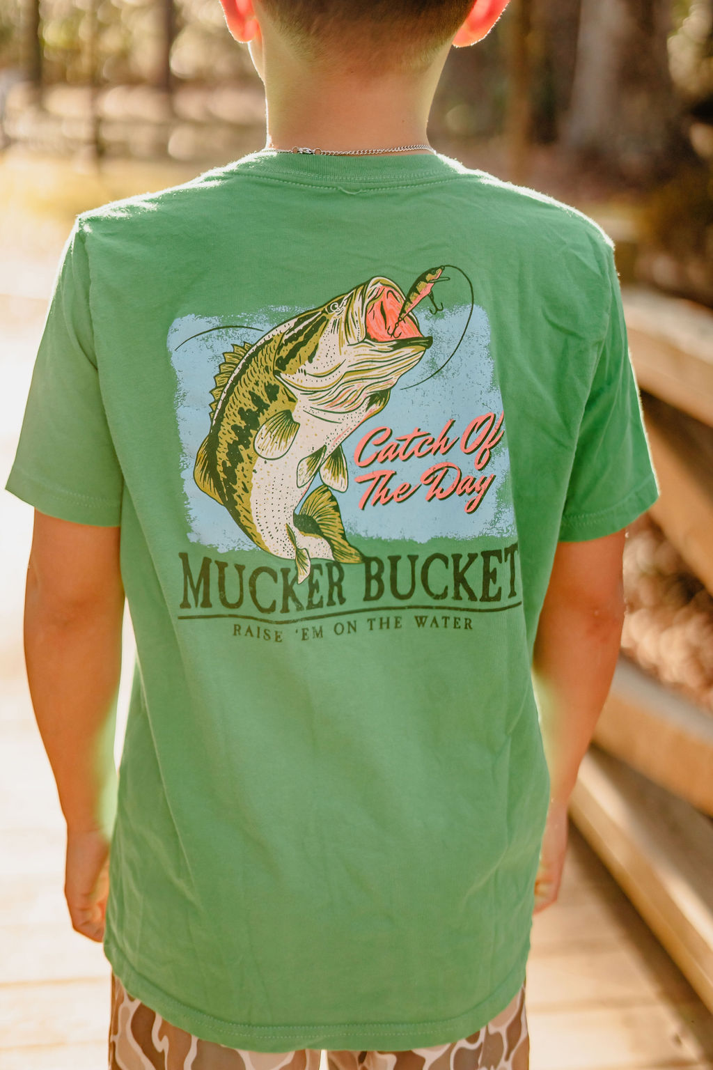 A child wears the Youth Catch of the Day tee by Mucker Bucket, boasting a vibrant fish graphic and the phrases Catch Of The Day and RAISE EM ON THE WATER. The charming sunlit outdoor setting with wooden planks enhances its appeal, ideal for youth.