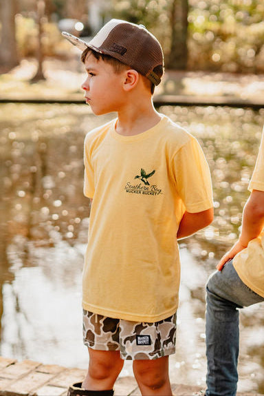 A young boy stands by a calm pond wearing a yellow T-shirt and Duck It Youth Performance Athletic Shorts by Mucker Bucket, looking to the side with hands behind his back. Sunlight filters through the trees, creating a peaceful outdoor setting.