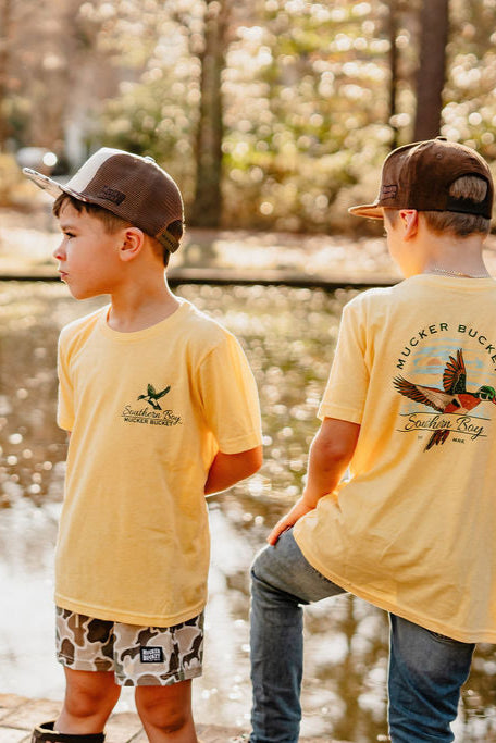 Two boys in Youth Southern Boy tees by Mucker Bucket stand by a pond, one facing the water and the other looking sideways. Sunlight filters through the trees, casting a warm, dappled effect on their yellow t-shirts and baseball caps.