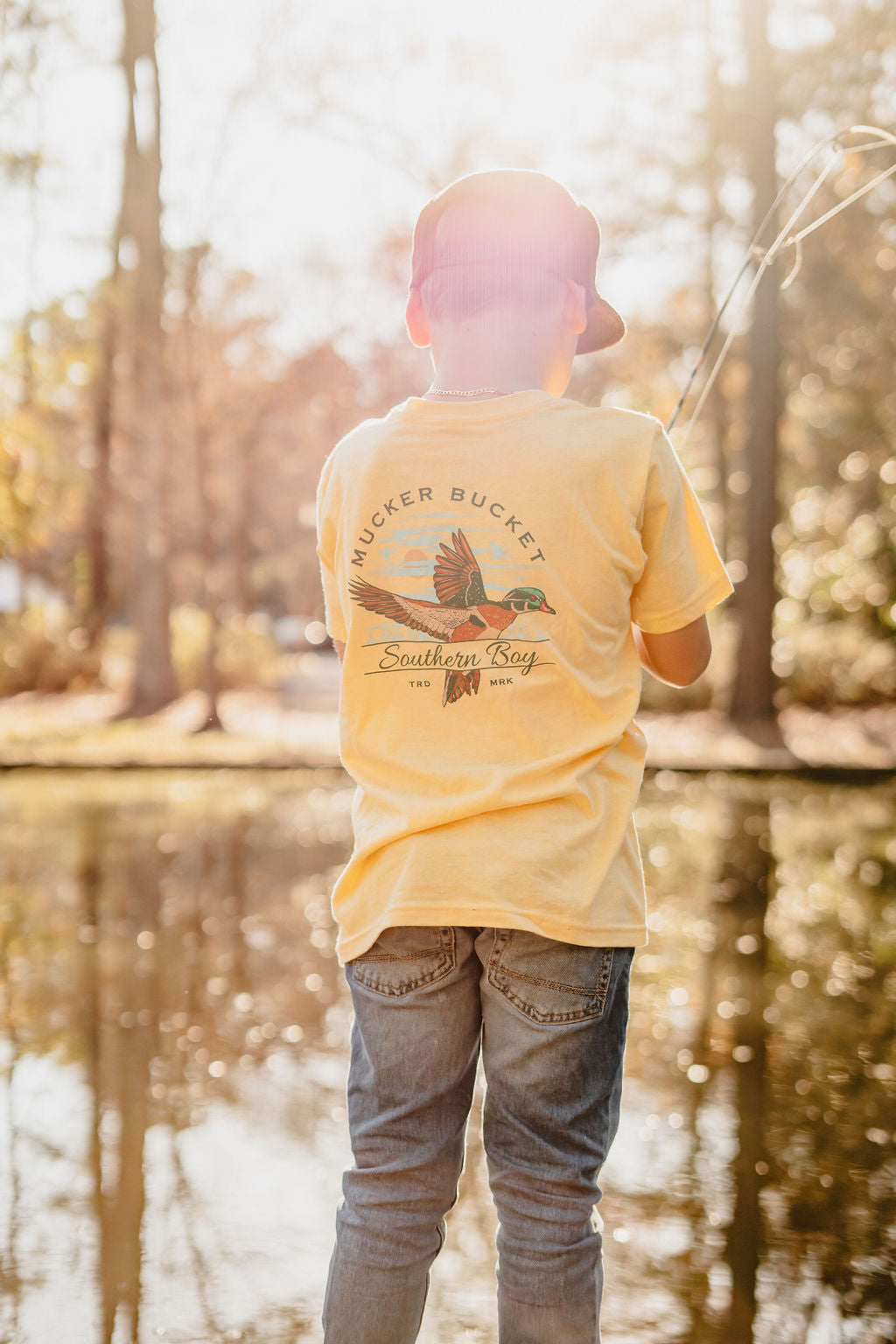 A child wearing a Mucker Buckets Youth Southern Boy tee, paired with a red cap and jeans, is fishing by a calm pond. The vibrant tee features a flying duck graphic, celebrating nature’s charm amidst the serene setting of sunlit trees in the background.
