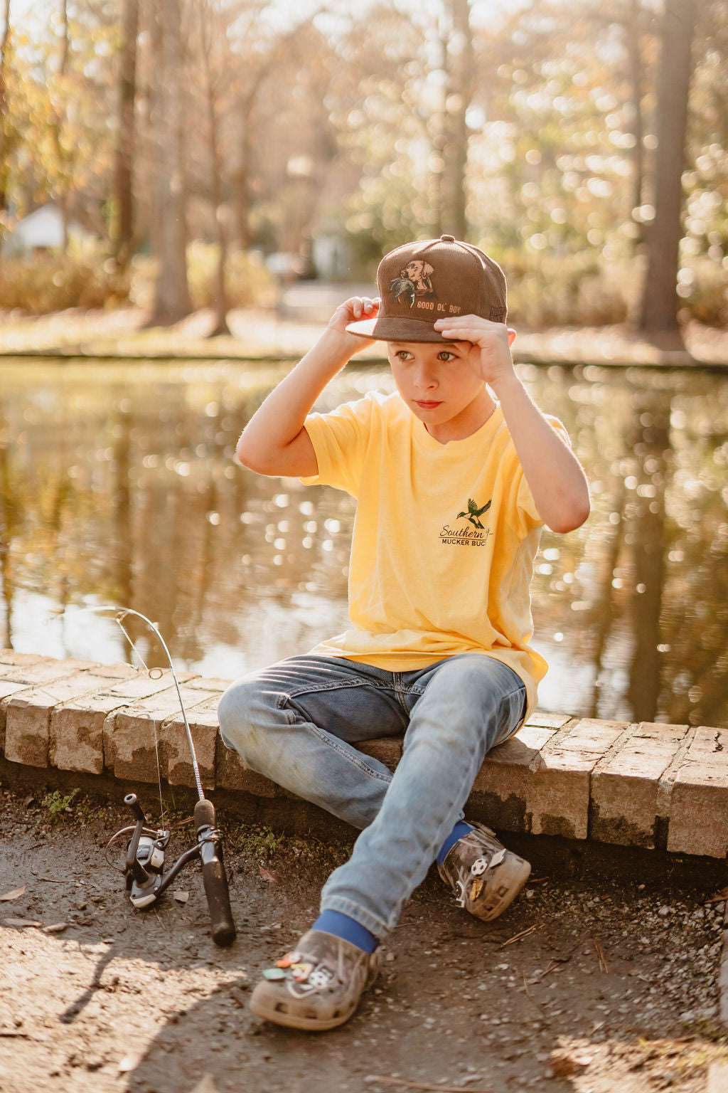 Wearing a Mucker Bucket Youth Southern Boy tee and jeans, a young boy sits by a pond on a sunny day, adjusting his cap. His fishing rod rests beside him as trees and warm lighting create an ideal backdrop for relaxing in youth-sized comfort.