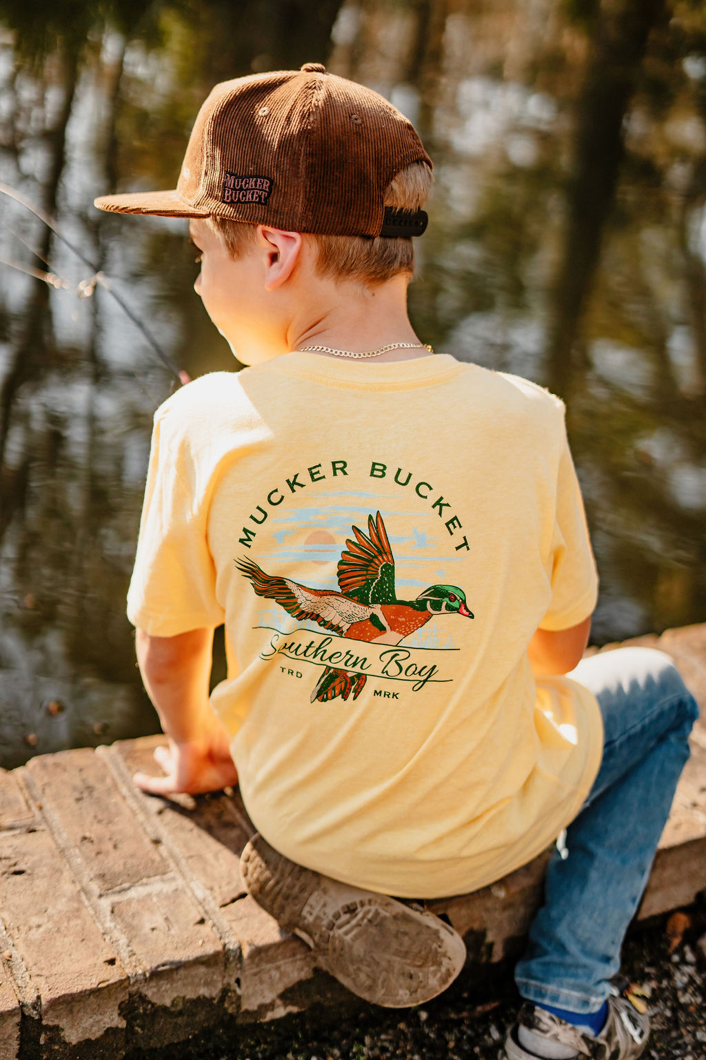 A young boy relaxes on a sunny brick ledge by the water, wearing a Youth Southern Boy T-shirt from Mucker Bucket. The yellow tee showcases a flying duck graphic with Mucker Bucket Southern Boy text, exuding a country vibe.