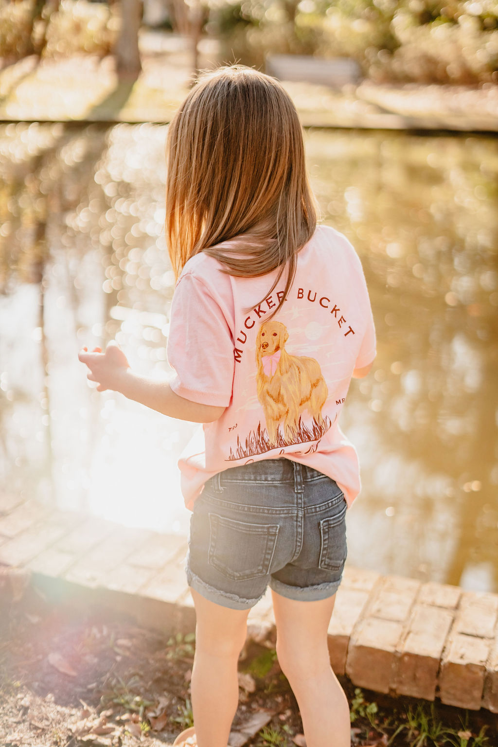 A child with long hair stands near a pond, wearing a pink Youth Southern Girl T-shirt by Mucker Bucket, featuring a cute dog graphic and denim shorts. Sunlight filters through the trees, highlighting the charm of this country-style tee in youth sizes.