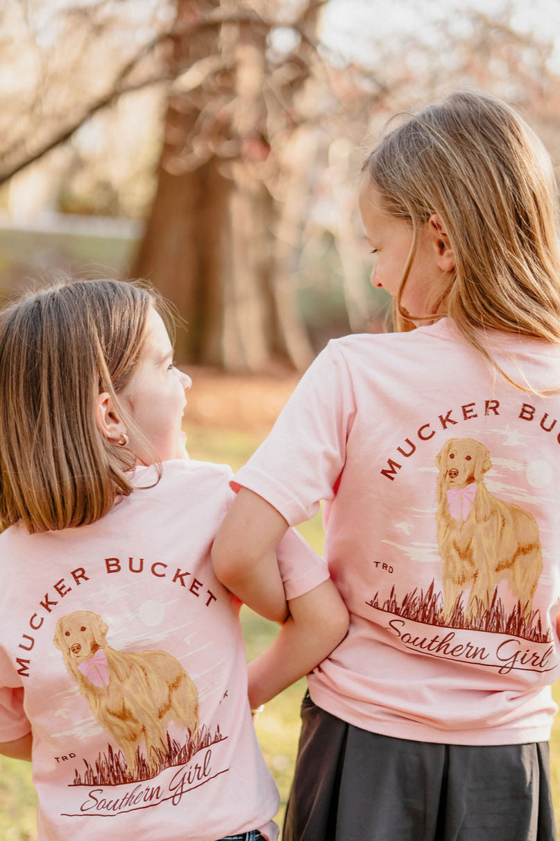 Two young girls outdoors in light pink, country-style tees featuring a cute golden retriever graphic. The Youth Southern Girl by Mucker Bucket is adorned with Mucker Bucket and Southern Girl on the back, as they smile at each other with trees in the background. Perfect for youth sizes!.