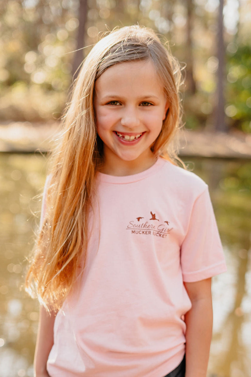 A young girl with long brown hair smiles near a pond, wearing the Youth Southern Girl t-shirt by Mucker Bucket. The light pink tee features a cute dog graphic and the words Southern Girl, set against trees and sunlight, enhancing its country style vibe.