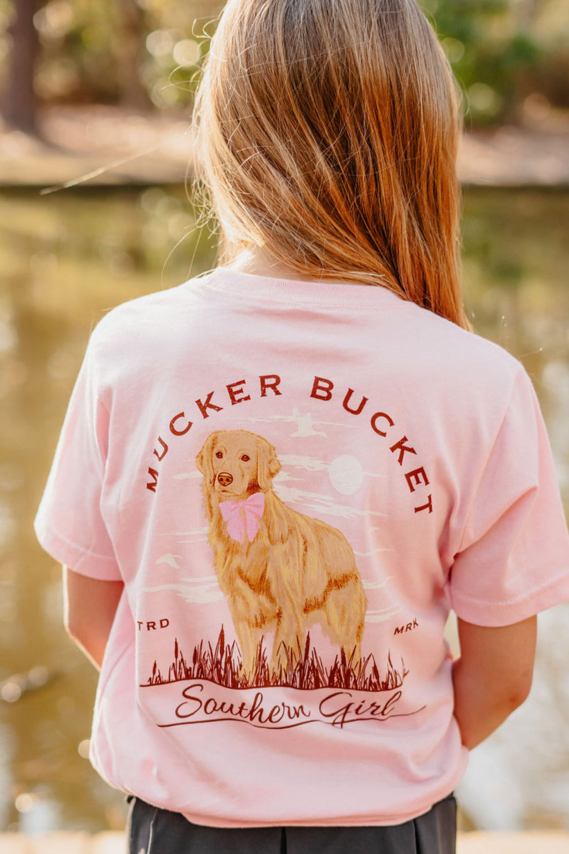 By a pond, a girl poses in the charming Youth Southern Girl tee from Mucker Bucket, featuring a graphic of a golden retriever with a pink bow tie. Her pink T-shirt reads MUCKER BUCKET and Southern Girl, perfectly complementing the serene scenery.